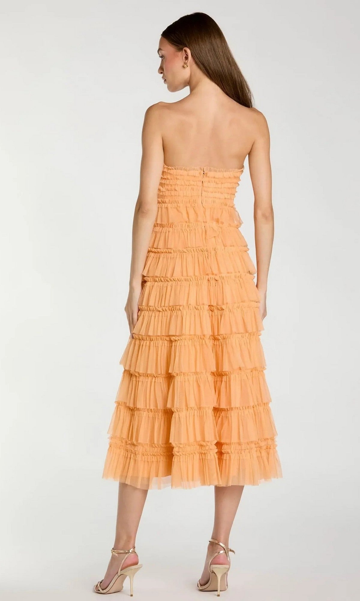 A woman stands facing away, wearing the Midi Semi-Formal Dress: Mac Duggal 8286—a strapless peach wedding guest dress with micro-ruffle detailing—paired with high-heeled sandals.